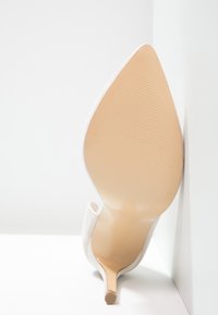Vit högklackad sko med spetsig tå och en slät, smidig yta. Klacken är tunn och stilletto-stil, vilande mot en vit yta.