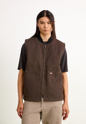 Brown sleeveless vest with a front zipper, ribbed collar, and two front pockets. Worn over a black shirt, paired with striped pants.
