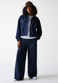 Veste en denim foncé avec deux poches avant, associée à un jean large foncé. T-shirt blanc visible. Casquette marron avec des accents au motif léopard.