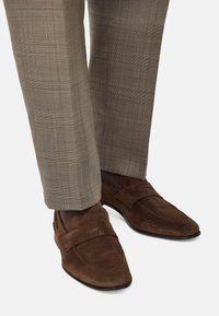 Brogādas krāsas zamšādas loafers ar krustenisku siksniņu detaļām, saskaņotas ar gaiši bēšiem kvadrātiem formētiem biksēm.