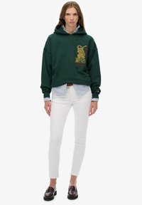 Vrouw draagt een forestgroene hoodie met tijgergrafiek, gecombineerd met een gestreept shirt, witte skinny broek en donkerbruine loafers, staand in een rechte houding.