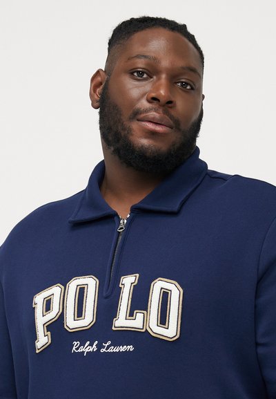 Marineblauwe fleece pullover met een ritssluiting bij de kraag, met "PO LO" in witte en gouden borduursel en "Ralph Lauren" eronder gestikt.