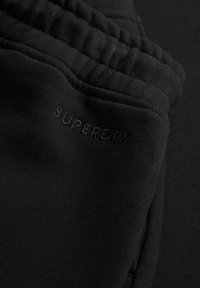 Close-up of black elastic waistband and pocket area on black Superdry sweatpants with embroidered brand name.