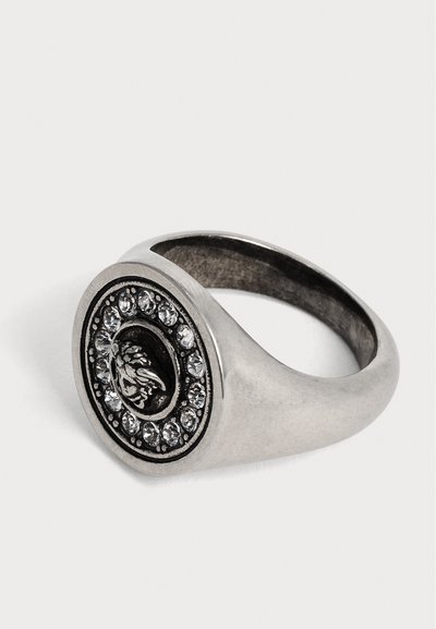Bague en argent avec un visage circulaire présentant un emblème noir au centre et un anneau de pierres précieuses claires autour.