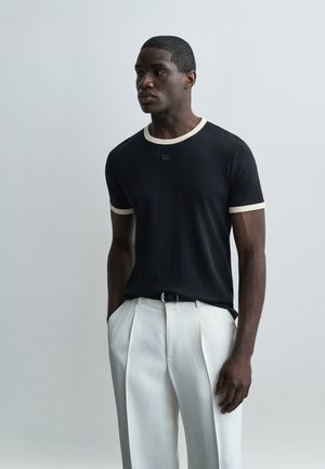 Homme portant un t-shirt noir avec des bordures blanches et un pantalon blanc plissé taille haute, debout devant un fond gris uni, les mains dans les poches.
