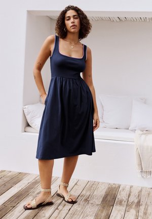 Göndör hajú nő, aki ujjatlan, térdig érő sötétkék ruhát és bézs szandált visel, fa padlón áll egy fehér ülőhely közelében.