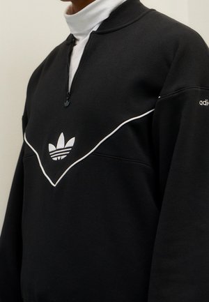 Osoba w czarnej bluzie Adidas z białym przeszyciem i logo, nałożonej na biały golf, widoczna od ramion do połowy tułowia.