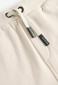 Helle beige Stoff-Jogginghosen mit einer Kordelzug-Schließe. Verfügt über schwarze Hardware und Etiketten mit dem Aufdruck "BAKER" in Weiß. Weiche Textur.