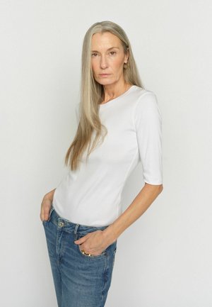 Mittelalte Frau mit langen grauen Haaren, die ein weißes eng anliegendes Oberteil und blaue Jeans trägt, mit den Händen in den Taschen stehend vor einem einfarbigen Hintergrund.