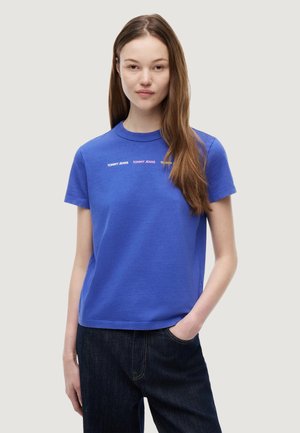 Joven mujer con el pelo largo y castaño, vestida con una camiseta azul de Tommy Jeans y vaqueros oscuros, de pie con una mano en el bolsillo.