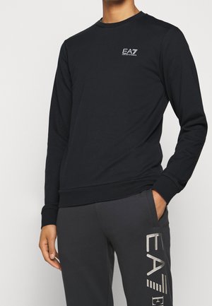 Mężczyzna w czarnej, długorękawowej bluzie EA7 Emporio Armani i ciemnoszarych spodniach dresowych z dużym logo EA7 na udzie, z ręką w kieszeni.