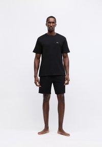 Homem em pé descalço, vestindo uma camisa preta de manga curta simples e calções pretos combinando, em fundo branco.
