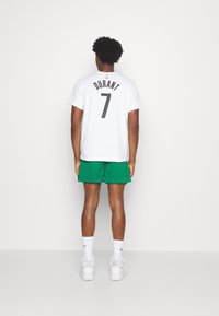 Uomo in piedi di spalle, che indossa una maglietta da basket bianca "Durant 7", pantaloni corti verdi, sneaker alte bianche e calze bianche Nike.