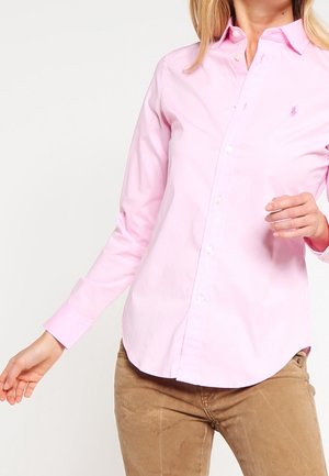 Vrouw die een lichtroze overhemd met lange mouwen en beige broek draagt, poseert met licht opgetilde armen.