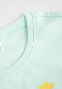 Zbliżenie na dekolt jasnolistnej zielonej bawełnianej koszulki z widocznym przeszyciem i częścią żółtego nadruku w kształcie gwiazd na tkaninie.