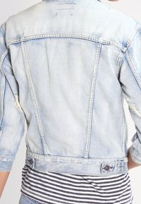 Light blue denim jacket featuring a faded design, distressed accents, and a button closure at the waist. Three-quarter sleeves visible.