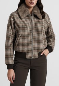 Donna che indossa una giacca a quadri con zip e colletto di pelliccia, con polsini e fascia in vita a coste di colore marrone scuro, abbinata a pantaloni scuri.