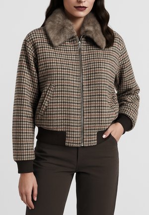 Donna che indossa una giacca a quadri con zip e colletto di pelliccia, con polsini e fascia in vita a coste di colore marrone scuro, abbinata a pantaloni scuri.