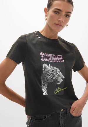 Femme portant un t-shirt noir avec un profil de léopard et le texte "SAVAGE" et "Lola Casademunt,", assorti à un jean foncé et des boucles d'oreilles créoles dorées.