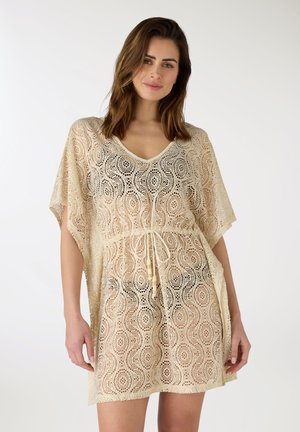 Donna con vestito copricostume beige all'uncinetto con cintura in vita, in piedi davanti a uno sfondo bianco semplice, rivolta in avanti.