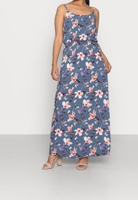 Kvinna i en blå maxikjol med rosa, lila och vita blommor och fjärilsmönster, kombinerad med beige klacksandaler.