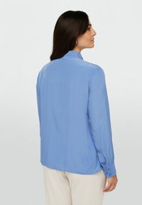 Blusa blu a maniche lunghe con una texture liscia, polsini con bottoni e collo alla cinese, caratterizzata da una vestibilità rilassata e dettagli con pieghe sul retro.
