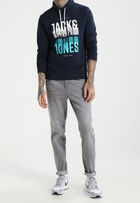 Felpa con cappuccio blu navy con stampa grafica in bianco e turchese, abbinata a jeans grigio chiaro con risvolti e sneakers bianche. Vestibilità casual e comoda.