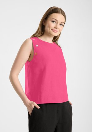 Vrouw draagt een mouwloze felroze top met kleine geborduurde motieven bij de schouder, gecombineerd met een zwarte broek, handen in de zakken.