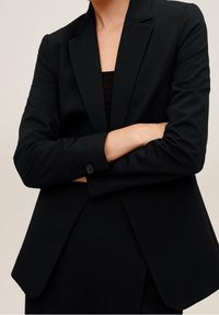 Blazer negro a medida con solapas en pico, mangas largas y puños abotonados. El tejido parece suave, con una forma estructurada y un diseño mínimo.