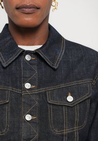 Jachetă din denim închis cu butoane metalice, cusături galbene și două buzunare frontale, având un guler clasic. Textură netedă, tăietură ajustată.