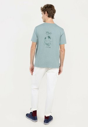 Joven de espaldas que lleva una camiseta gráfica azul claro, pantalones blancos, calcetines granate y zapatillas azules sobre un fondo liso.