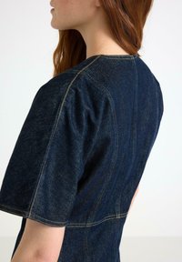 Mörkblå denimtop med rundad halsringning, korta puffärmar och synlig gul sömnad, som visar en figurnära design och strukturerade sömmar.