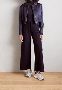 Mörkblå sidenjacka i kort modell över en figurnära svart polotröja, ihop med svarta vida byxor och silvriga sportiga sneakers.