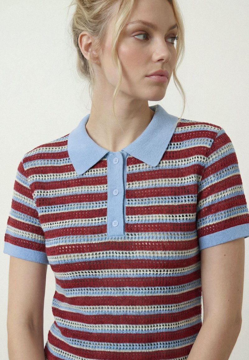 Femme aux cheveux blonds attachés en arrière portant un polo en maille à manches courtes avec des rayures horizontales rouges, bleues, blanches et beige, regardant sur le côté.