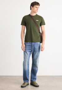 Ελαιόχρωμο βαμβακερό t-shirt, μπλε τζιν παντελόνι και ελαιόχρωμα αθλητικά παπούτσια. Διαθέτει λογότυπο στο μπλουζάκι και καφέ χιαστί λουρί.