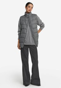 Grå vadderad väst med hög krage och tryckknappsstängning, bärs över en grå topp och parkeras med vida mörkgrå byxor. Slät textur, avslappnad design.