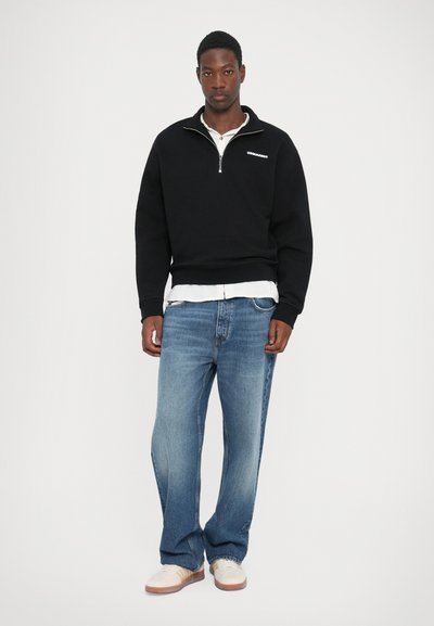 Zwarte rits-sweater met ribgebreide zoom, gelaagd over een wit shirt. Gepaard met lichtblauwe rechte spijkerbroek en lichtgekleurde sneakers.