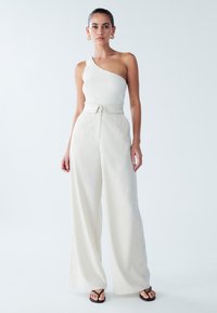 Weißes, geripptes One-Shoulder-Oberteil kombiniert mit hellbeigen, weit geschnittenen Hosen. Die Hose hat eine taillierte Passform mit Gürtel und eine weiche Textur. Schwarze Flip-Flops.