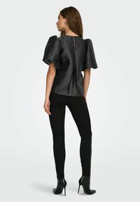 Mujer vestida con blusa negra de satén con mangas abullonadas, pantalones negros ajustados con adornos laterales y botas de tacón stiletto negras hasta el tobillo, de espaldas.