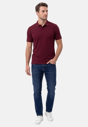 Mand iført en burgundy kortærmet polo, blå jeans, hvide sneakers, stående med den ene hånd i lommen og kigger mod højre.