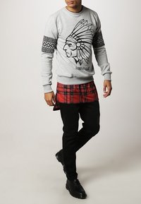 Gray sweatshirt with a feathered headdress graphic, black accents on the sleeves, layered over a red plaid shirt, paired with black pants.