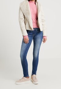 Vit jacka fodrad med fårskinn, rosa tröja, blå jeans och beige sneakers med texturerad yta och snördetaljer. Avslappnad outfitkombination.