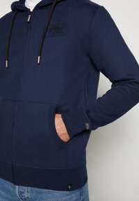 Navy Zip-Hoodie aus weichem Stoff, mit schwarzen Kordeln, Fronttaschen, gerippten Bündchen und einem kleinen Logo-Patch am Saum.