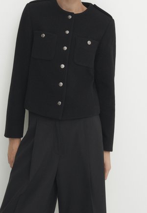 Kobieta ubrana w czarną krótko skrojoną kurtkę z srebrnymi guzikami i dwoma przednimi kieszeniami, w połączeniu z czarnymi szerokimi spodniami z wysokim stanem.