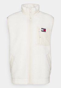 Beżowa kamizelka polarowa z wysokim kołnierzem, zamkiem błyskawicznym z przodu i kieszenią na piersi z naszywką z logo. Z boku znajdują się kieszenie.