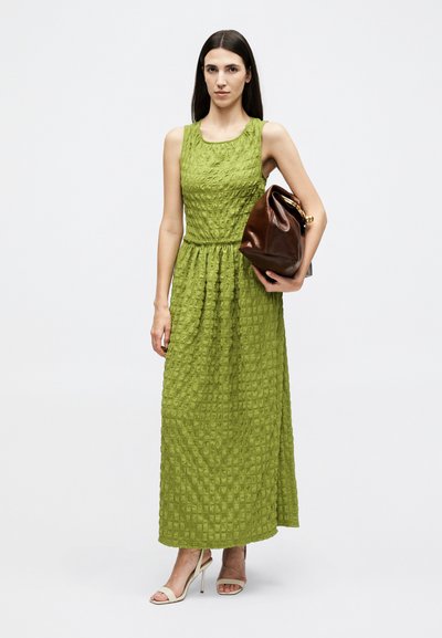 Γυναίκα που φοράει υφασμάτινο πράσινο αμάνικο μάξι φόρεμα, κρατώντας καφέ δερμάτινη τσάντα και φορώντας μπεζ τακούνια.