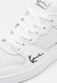 White sneaker with perforated leather upper, thick white laces, signature detail in navy, and a textured sole design.