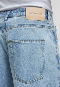 Jeans de mezclilla azul claro con un acabado texturizado, que cuentan con una etiqueta de marca de cuero beige y dos bolsillos traseros con costuras visibles.