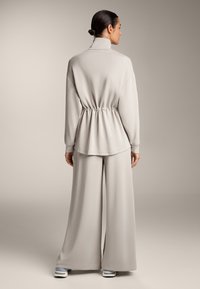 Lichtgrijze, lange mouwen pullover met een hoge kraag en een gerimpelde taille, gecombineerd met een broek met wijde pijpen. Glad materiaal met een ontspannen pasvorm.