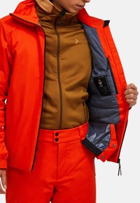 Lys orange vandtæt jakke over et brunt inderlag. Jakken har en lynlåslukning foran, en kontrastfuld blå foring og lomme.